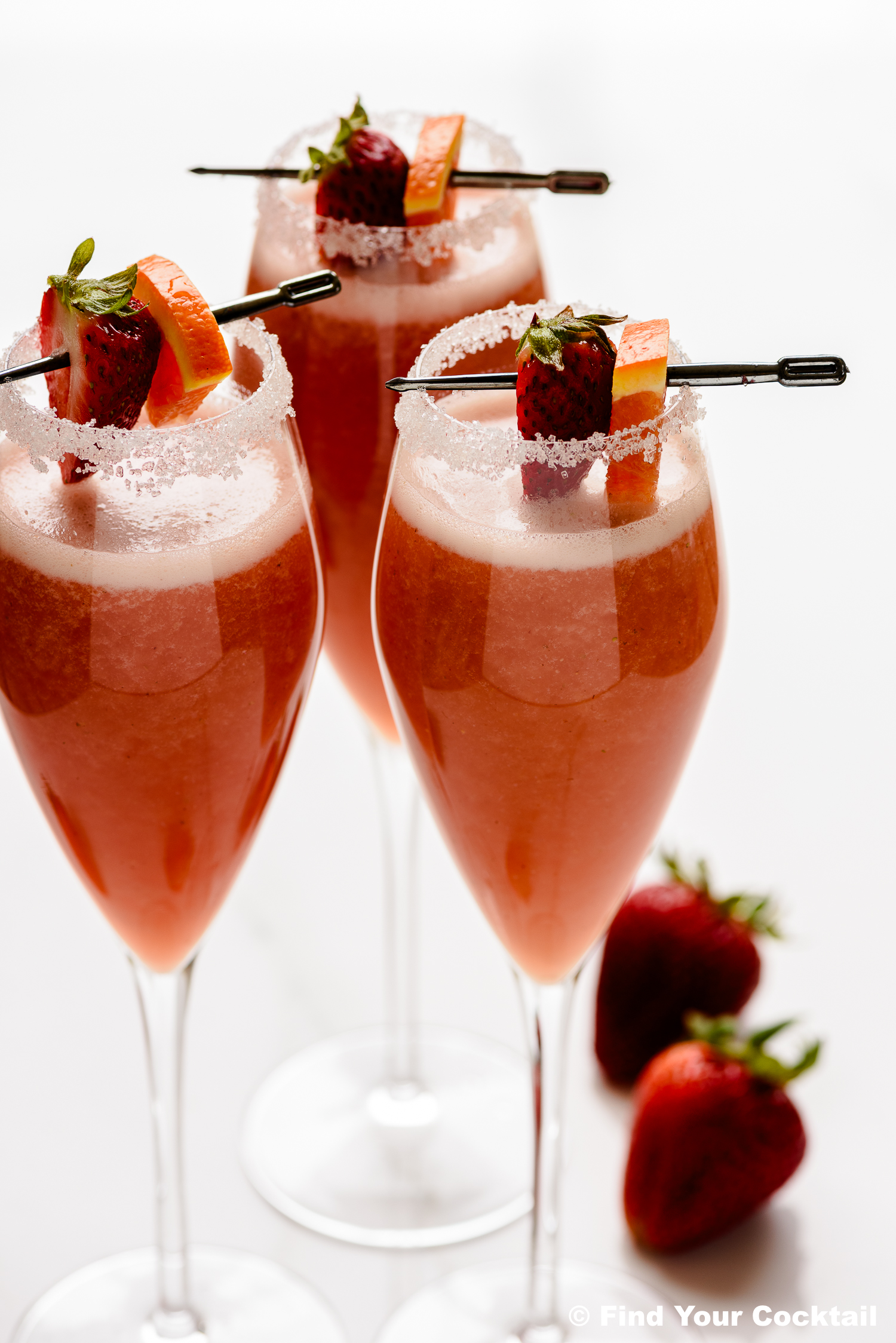 Three strawberry cocktails in flute glasses with sugar rims, garnished with strawberry and orange slices on skewers. Two whole strawberries are placed beside the glasses.
