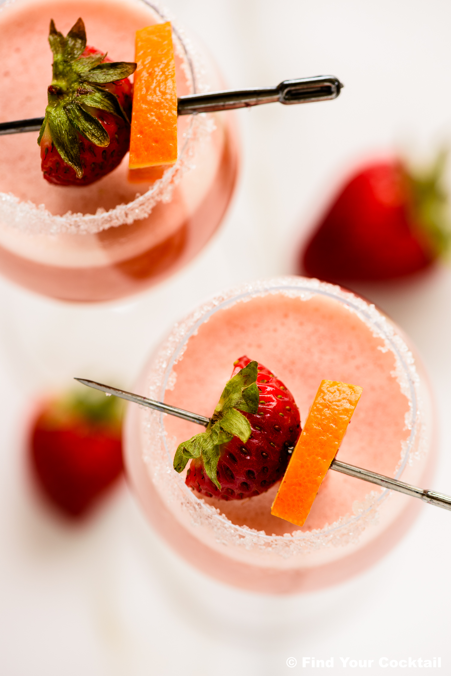 Two pink cocktails in sugar-rimmed glasses, each garnished with a whole strawberry and an orange slice on a skewer. Whole strawberries are placed nearby on a white surface.