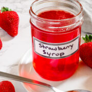 A glass jar labeled Strawberry Syrup sits on a white plate with a spoonful of syrup, surrounded by fresh strawberries on a marble surface.
