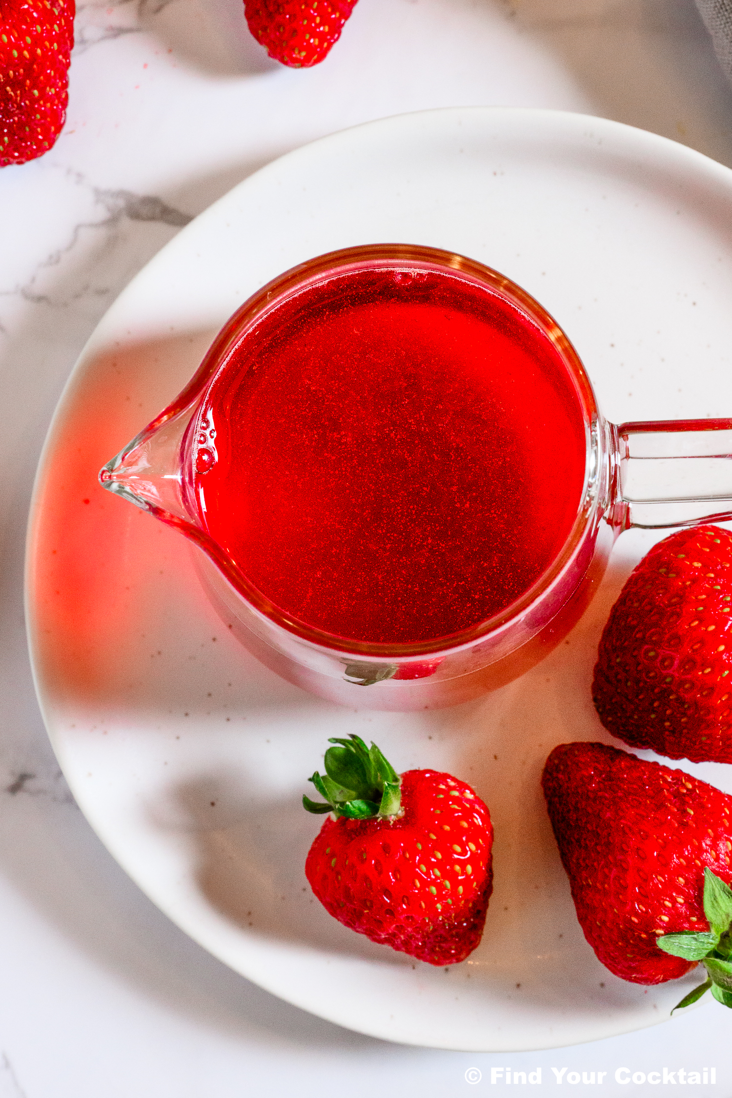 A glass pitcher filled with red liquid sits on a white plate, surrounded by fresh strawberries on a marble surface.