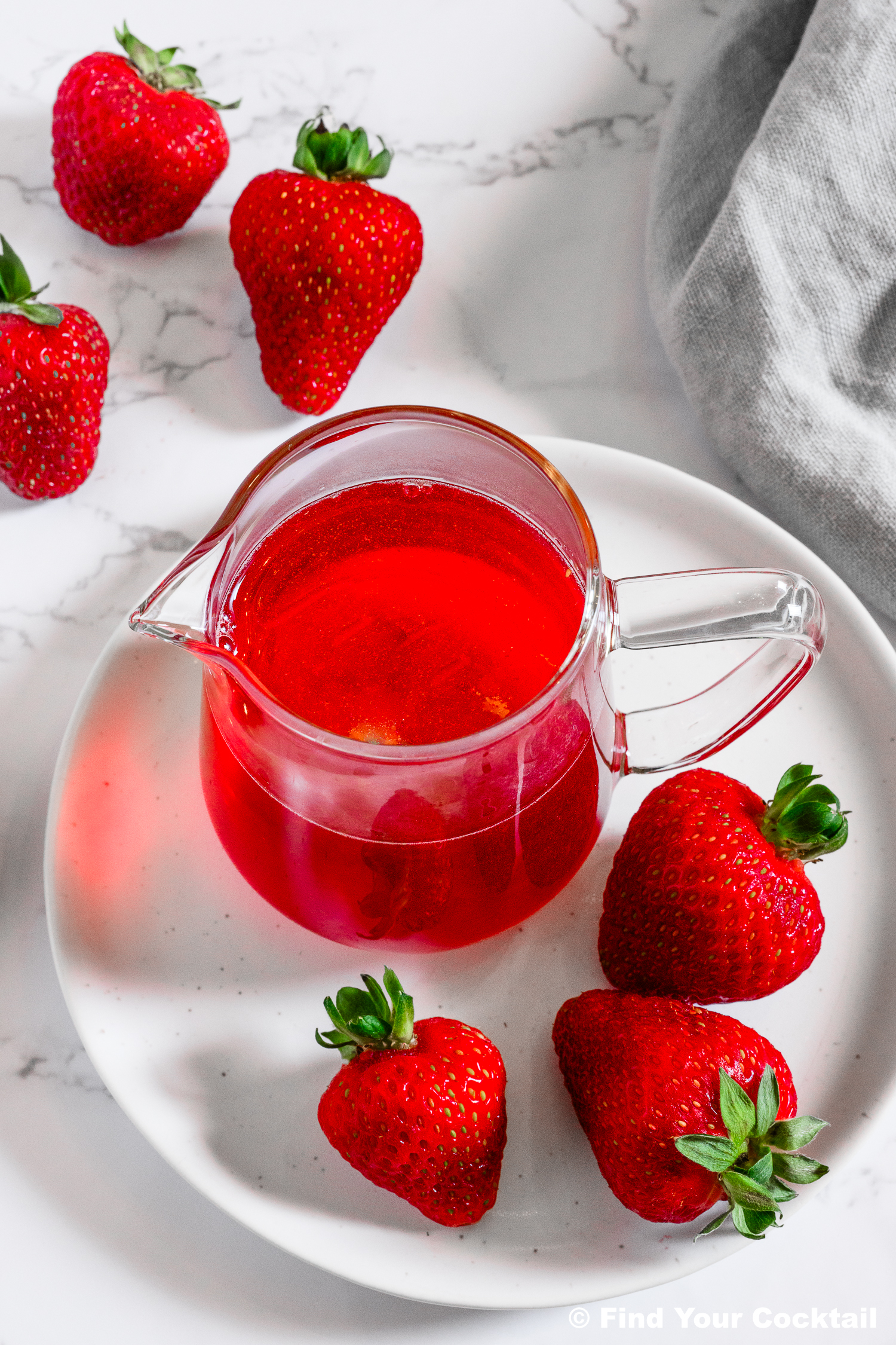 A clear glass pitcher of red liquid on a white plate with fresh strawberries, with more strawberries placed around the plate on a marble surface.