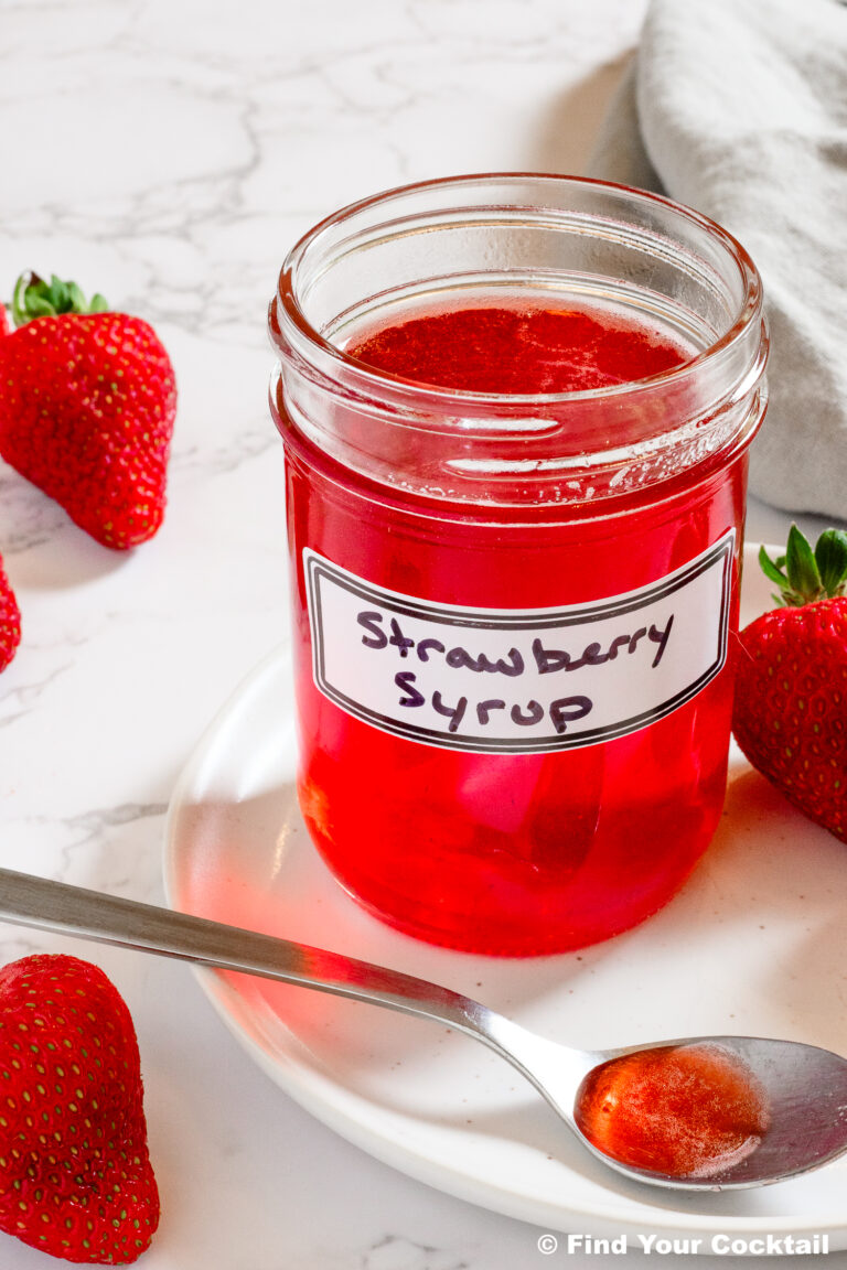 A glass jar labeled Strawberry Syrup sits on a white plate with a spoonful of syrup, surrounded by fresh strawberries on a marble surface.