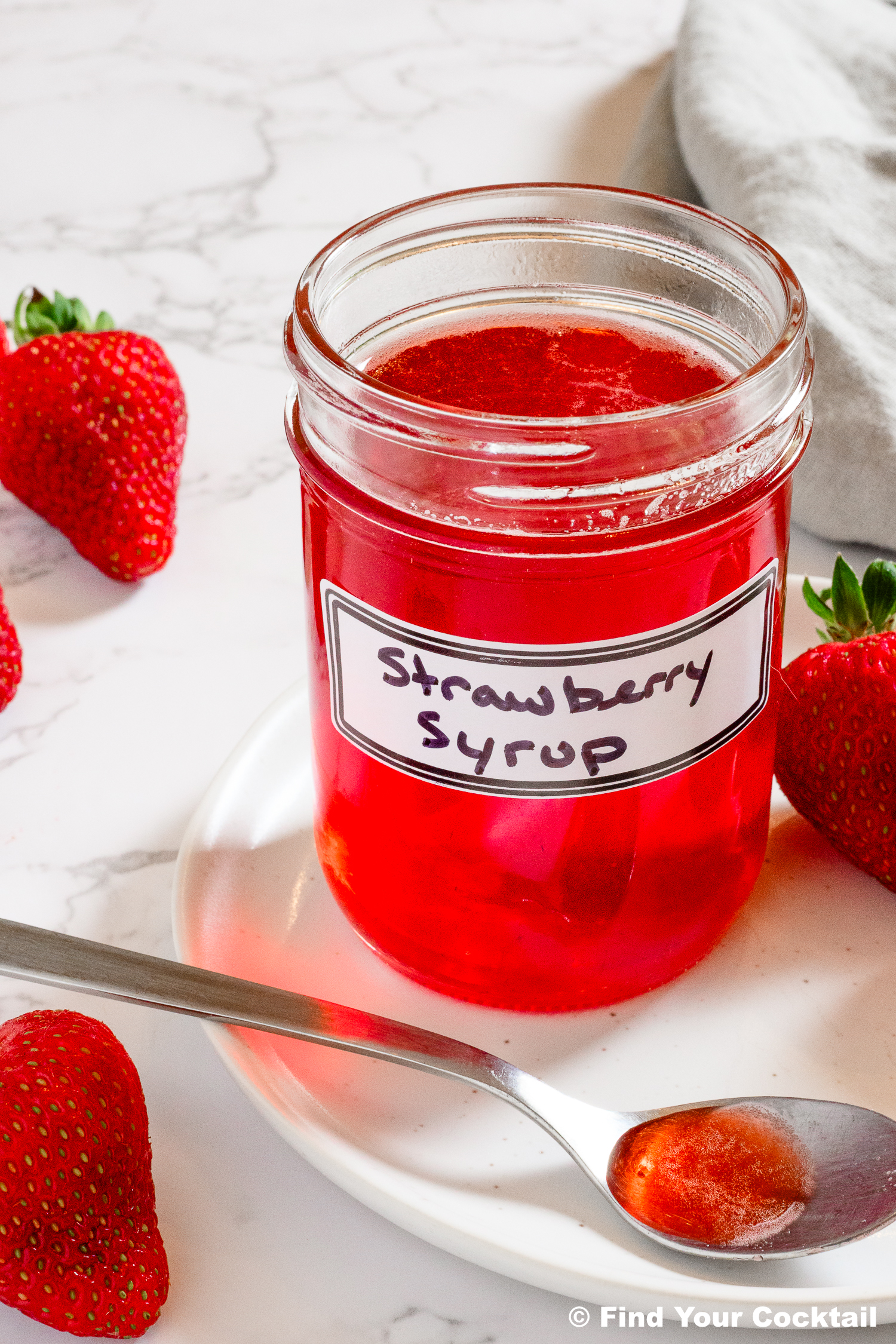A glass jar labeled Strawberry Syrup sits on a white plate with a spoonful of syrup, surrounded by fresh strawberries on a marble surface.
