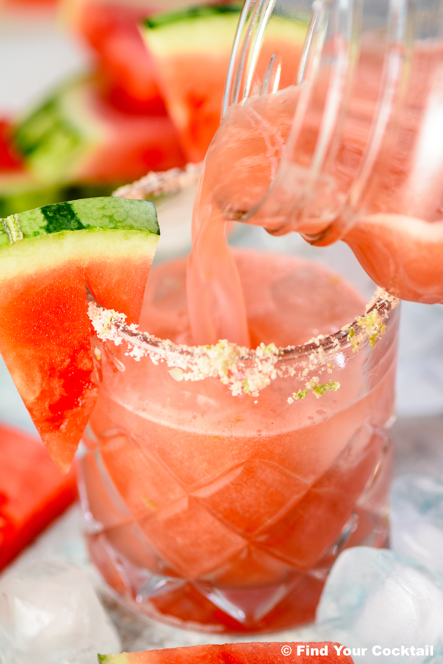 A glass rimmed with salt and garnished with a watermelon slice is being filled with a pink beverage, surrounded by ice and watermelon pieces.
