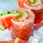 Two glasses of watermelon cocktail with salted rims, garnished with watermelon slices, lime, and jalape&ntilde;o, surrounded by ice cubes and watermelon pieces.