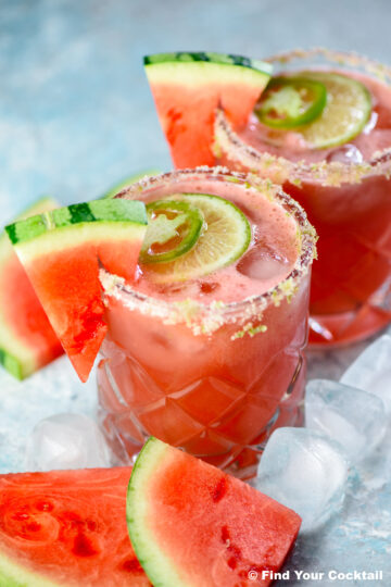 Two glasses of watermelon cocktail with salted rims, garnished with watermelon slices, lime, and jalapeño, surrounded by ice cubes and watermelon pieces.