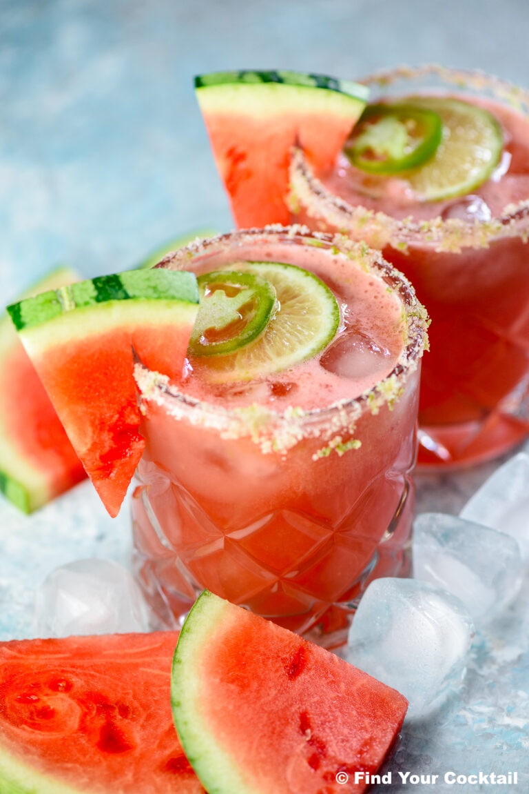 Two glasses of watermelon cocktail with salted rims, garnished with watermelon slices, lime, and jalapeño, surrounded by ice cubes and watermelon pieces.