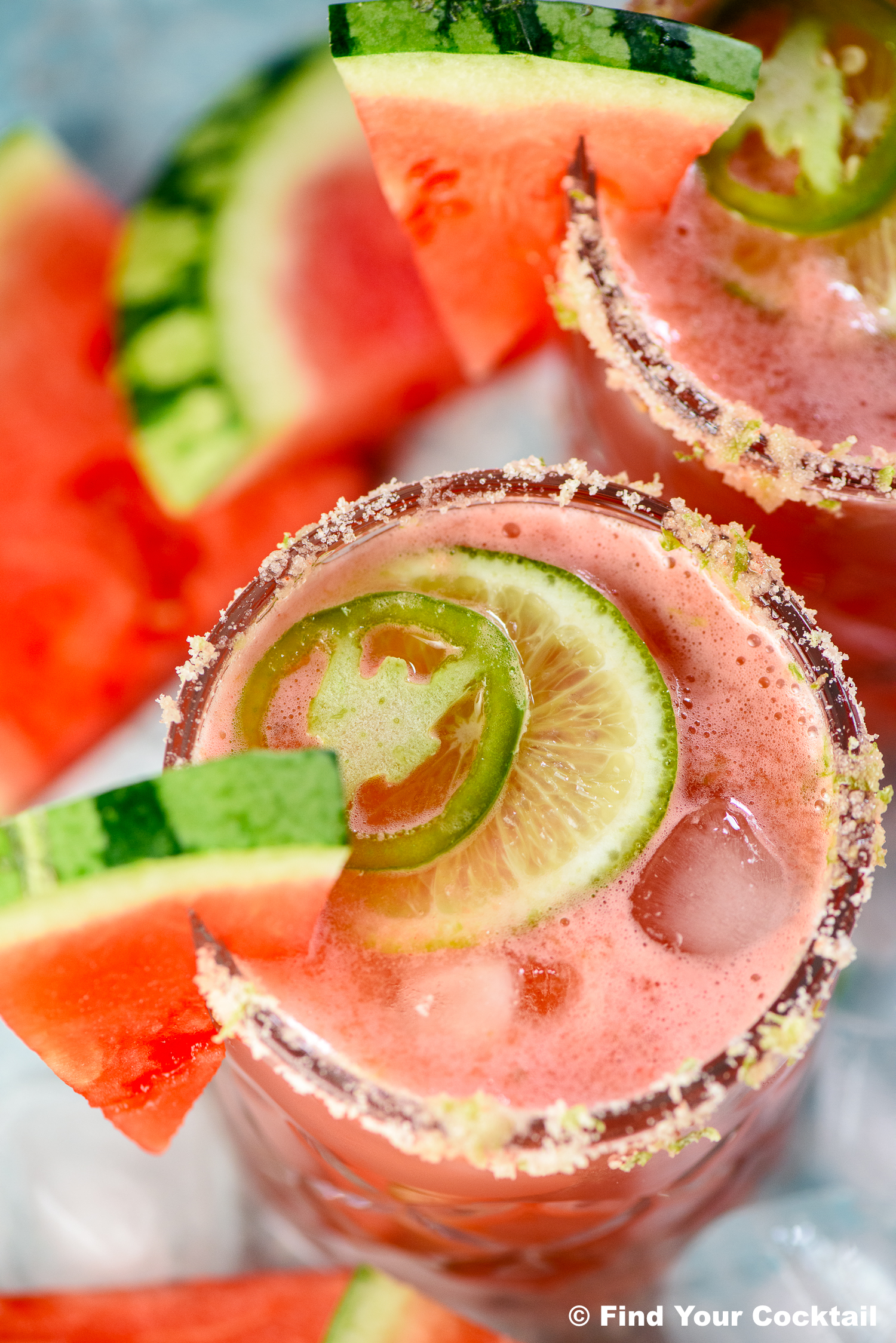A close-up of a pink cocktail with ice, garnished with lime, jalapeño, and a watermelon wedge, served in a glass with a seasoned rim.