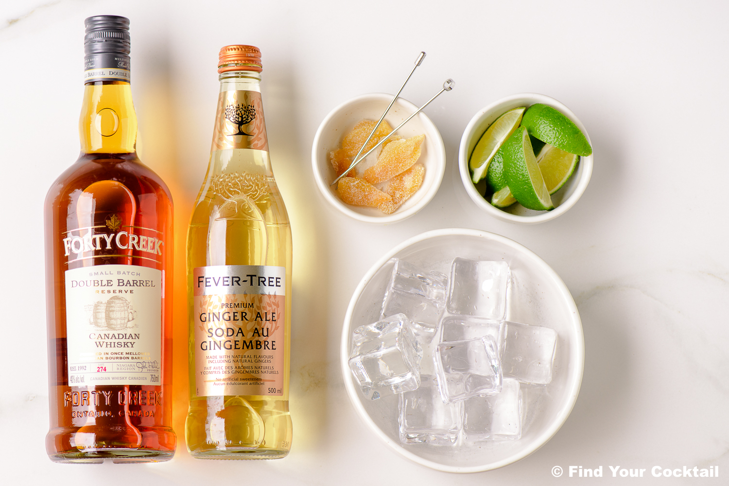 A bottle of Forty Creek Canadian Whisky, a bottle of Fever-Tree Ginger Ale, a bowl of ice cubes, a bowl of lime wedges, and a bowl of candied ginger on a white surface.