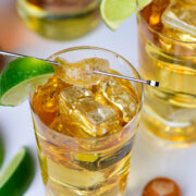 Two glasses of golden cocktail with ice, each garnished with a lime wedge and sugared rim, sit on a white surface near lime slices and a bottle cap.