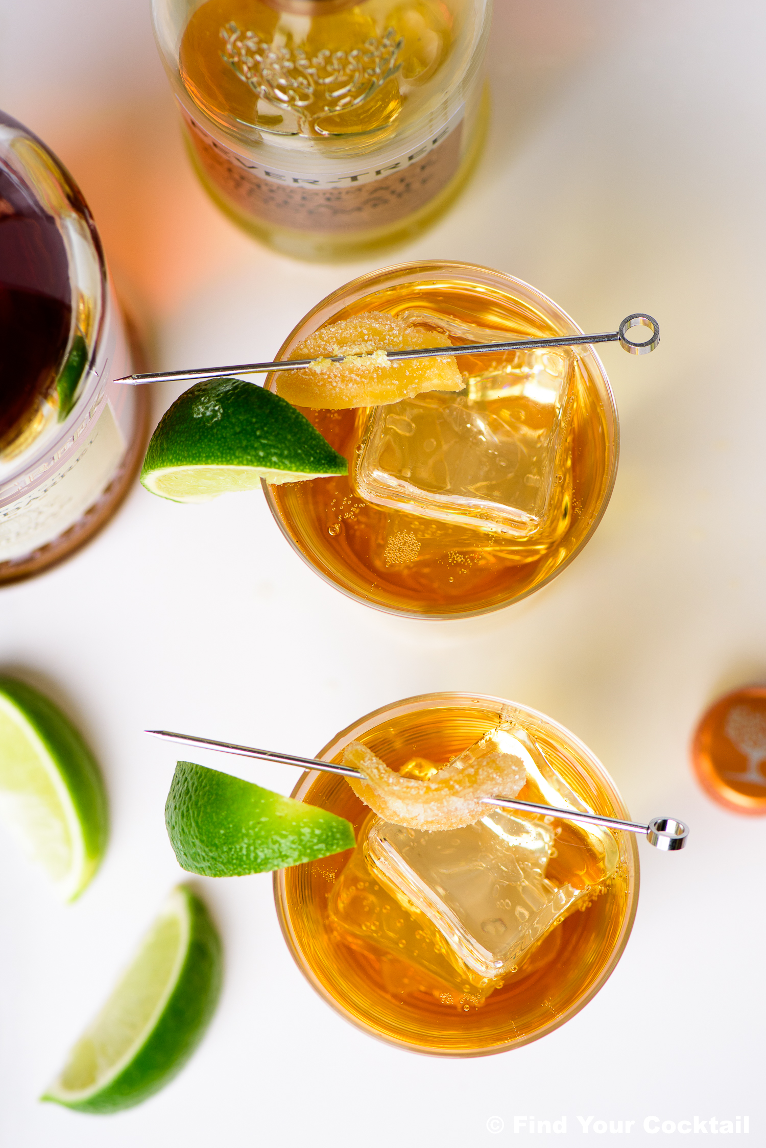 Two glasses of amber-colored cocktails with ice, garnished with lime wedges and cocktail picks, are placed on a white surface with liquor bottles and extra lime wedges nearby.