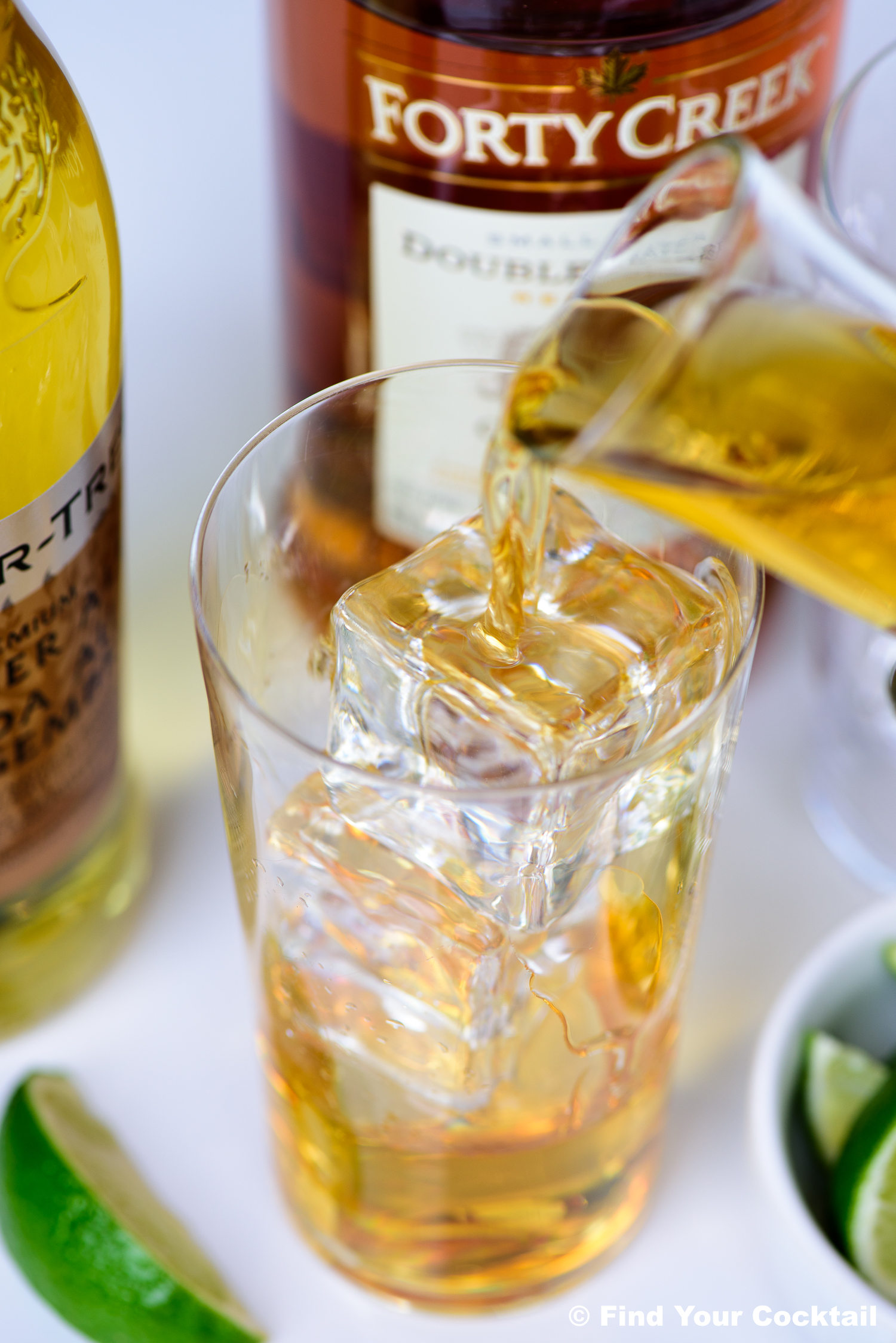 Whiskey is being poured over ice cubes into a glass, with whiskey bottles and lime wedges visible in the background.