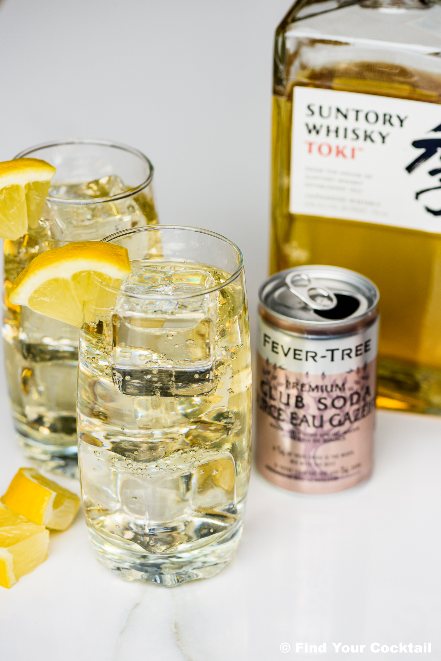 Two highball glasses with whisky, soda, ice, and lemon wedges, next to a bottle of Suntory Whisky Toki and a can of Fever-Tree club soda on a white surface.