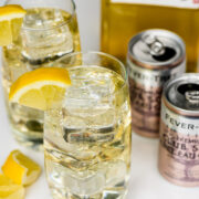 Two highball glasses with ice and lemon wedges, filled with a clear drink, are placed next to a bottle of Suntory whisky and two cans of Fever-Tree club soda.