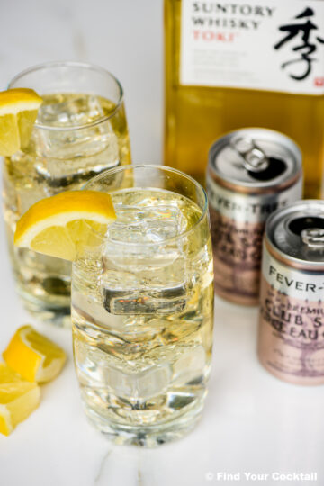 Two highball glasses with ice and lemon wedges, filled with a clear drink, are placed next to a bottle of Suntory whisky and two cans of Fever-Tree club soda.