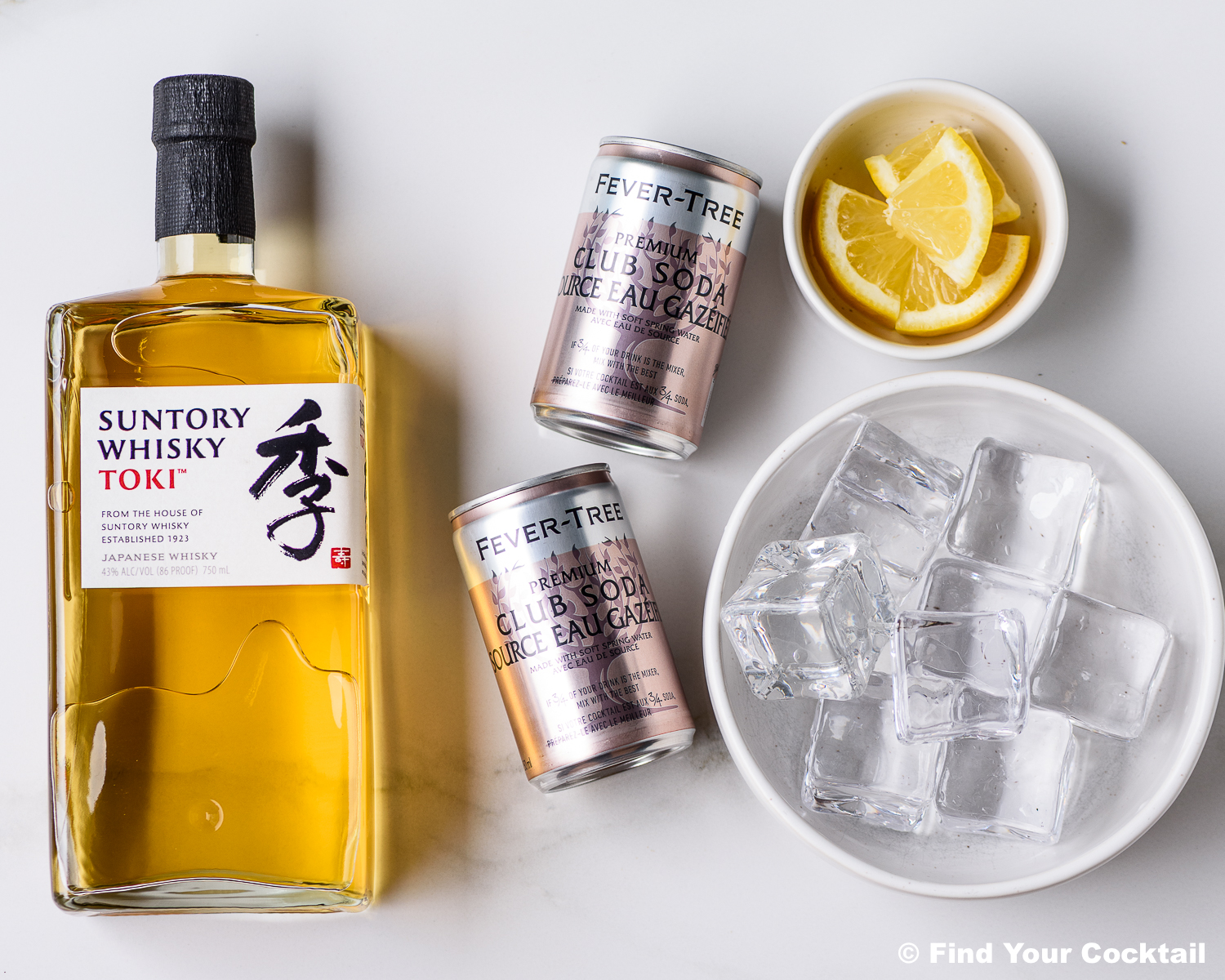 A bottle of Suntory Whisky Toki, two cans of Fever-Tree club soda, a bowl of lemon wedges, and a bowl of ice cubes on a white surface.