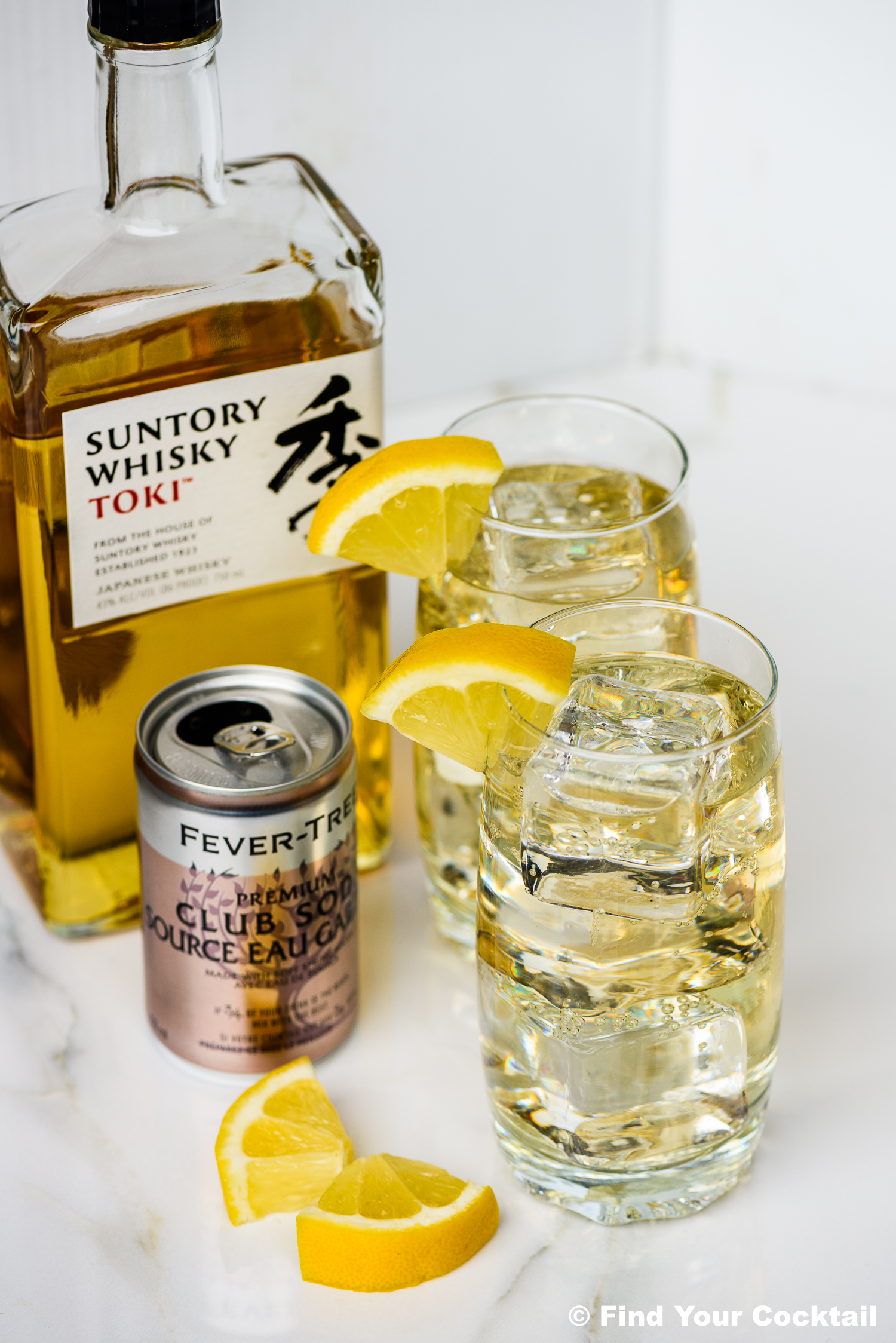 A bottle of Suntory Whisky Toki, a can of Fever-Tree club soda, two highball glasses with ice, whisky, soda, and lemon wedges, plus extra lemon slices on a white surface.