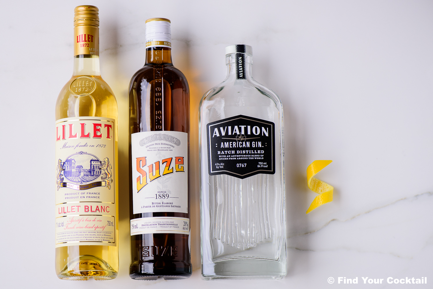Three bottles—Lillet Blanc, Suze, and Aviation Gin—stand side by side on a white surface next to a lemon twist.