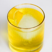 A clear glass with a yellow cocktail, a large square ice cube, and a lemon twist garnish, set on a white surface.