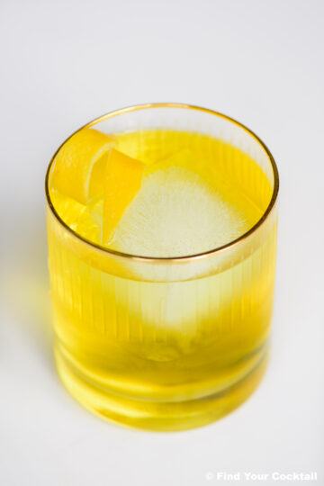 A clear glass with a yellow cocktail, a large square ice cube, and a lemon twist garnish, set on a white surface.