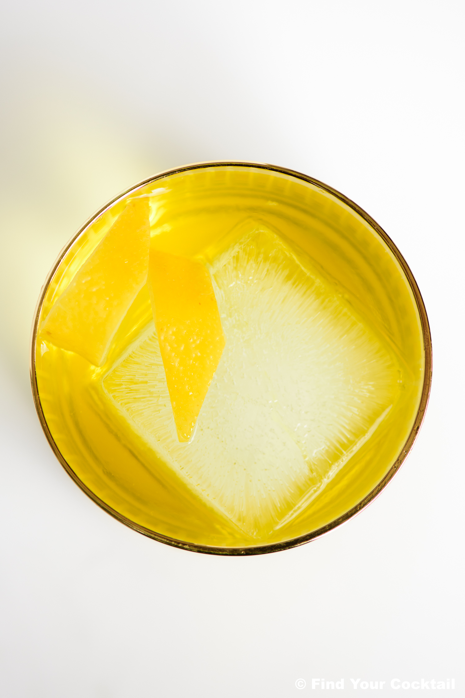 A yellow cocktail with a large clear ice cube and two lemon peels, served in a round glass, viewed from above.