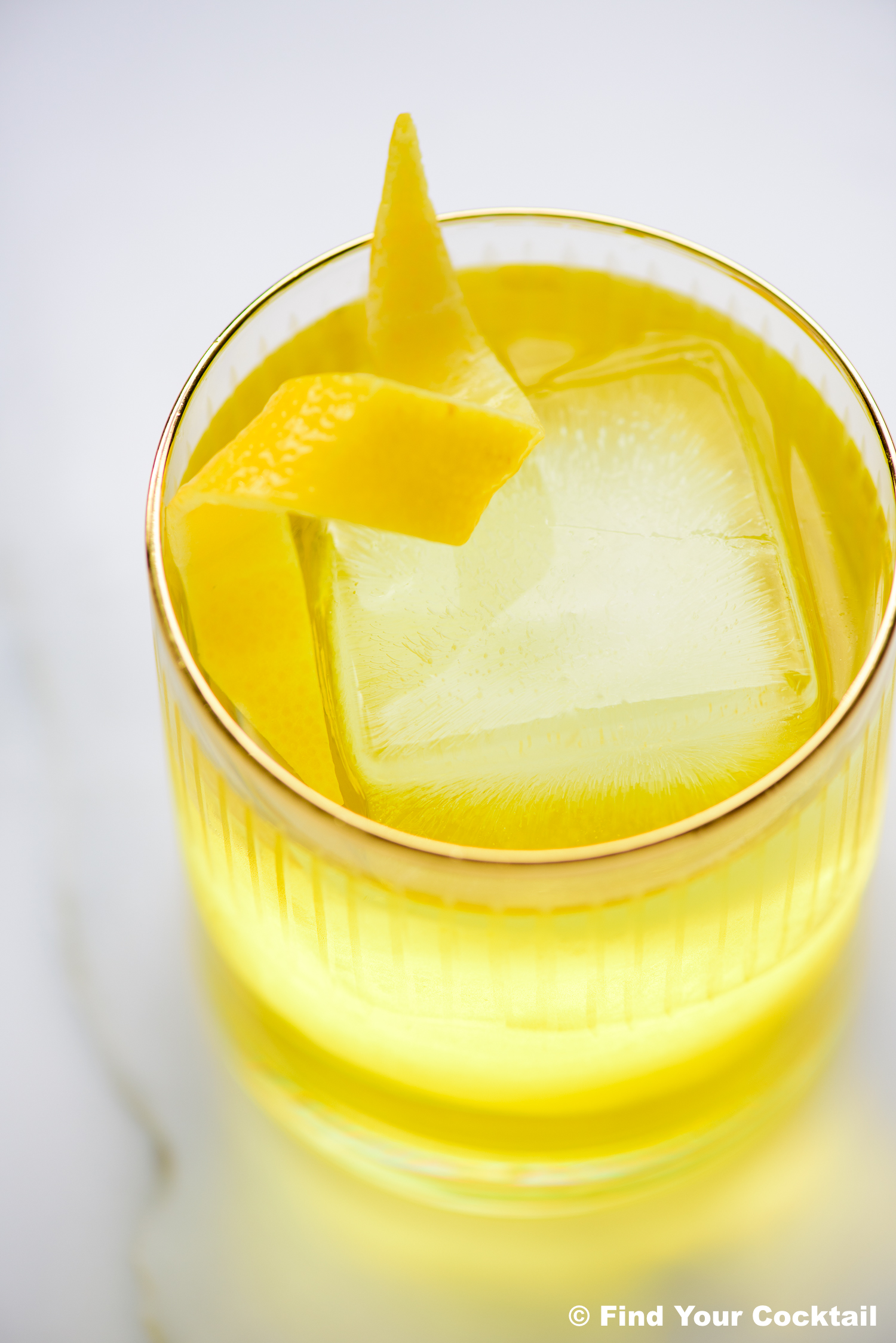 A clear glass filled with a yellow cocktail, large square ice cube, and a lemon twist garnish, placed on a white surface.