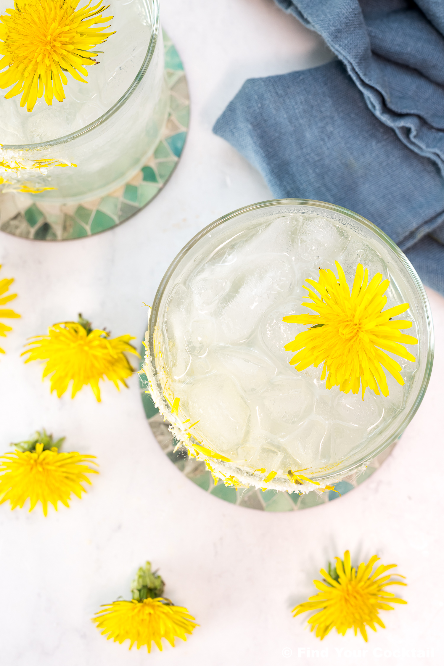 Overhead view of two glasses filled with ice and a clear drink, each garnished with a yellow dandelion flower, with additional dandelions and a blue napkin nearby.