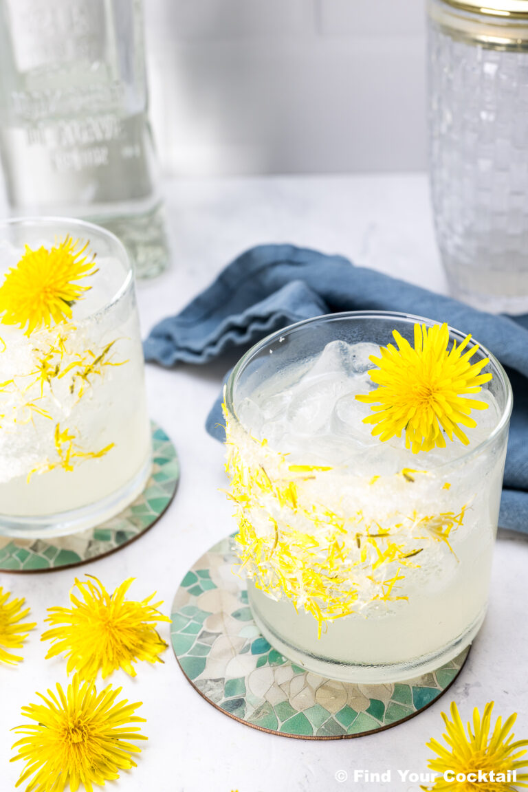 Two glasses of iced cocktails garnished with yellow dandelion flowers sit on patterned coasters, with a blue napkin and a bottle in the background.