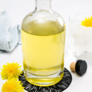 A clear glass bottle filled with yellow liquid is placed on a black coaster beside two dandelion flowers, a bottle cap, and a glass with ice and a dandelion flower.