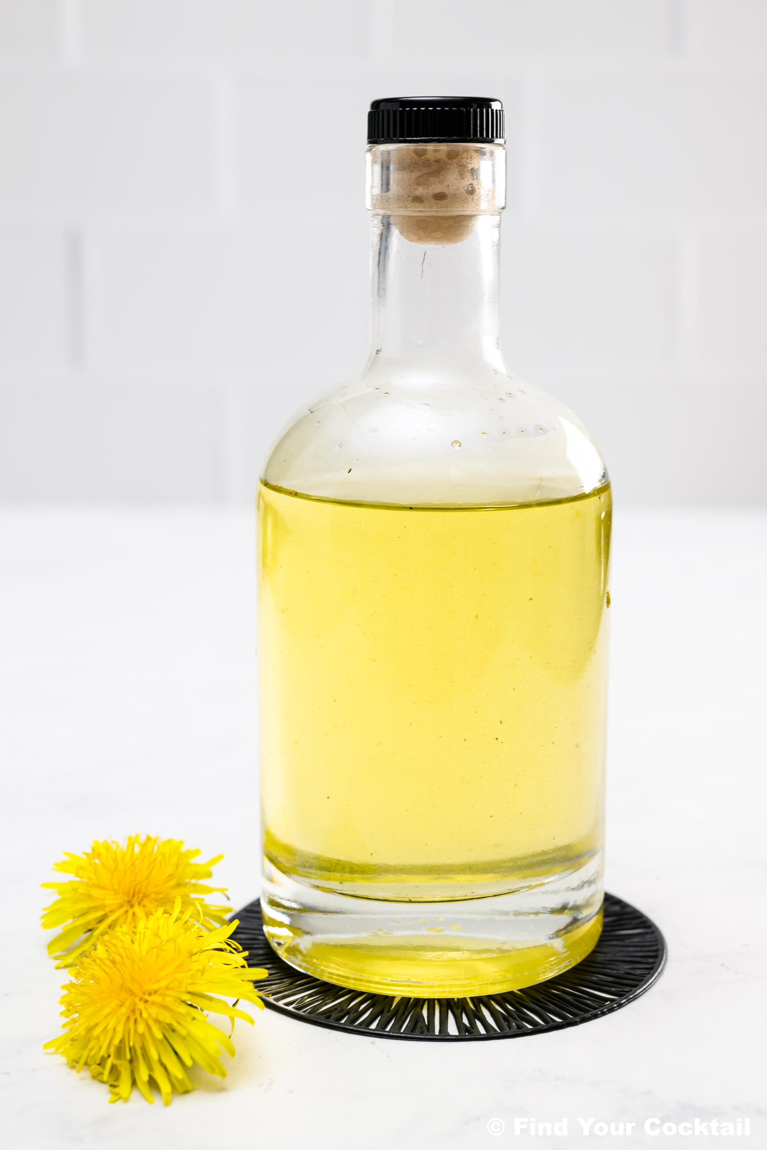 A clear glass bottle filled with yellow liquid, sealed with a black cap and cork, sits on a coaster next to two yellow dandelion flowers.