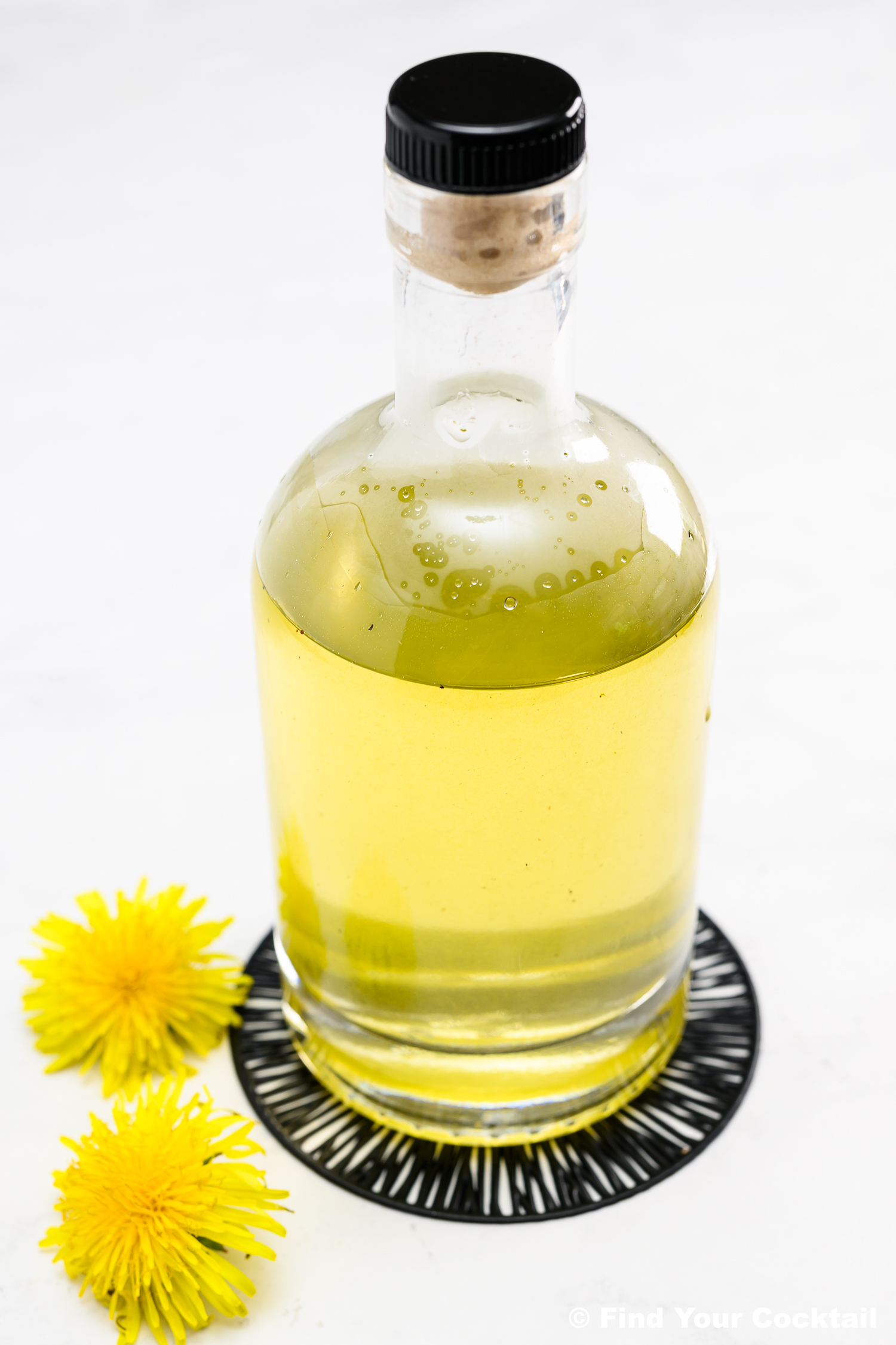 A clear glass bottle filled with yellow liquid sits on a round black and white coaster, with two yellow dandelion flowers beside it on a white surface.
