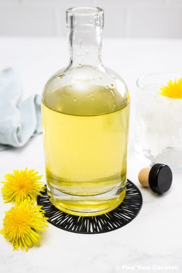 A clear glass bottle filled with yellow liquid is placed on a black coaster beside two dandelion flowers, a bottle cap, and a glass with ice and a dandelion flower.