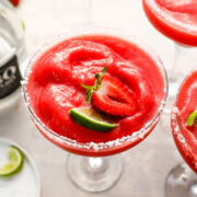 A frozen strawberry margarita in a salt-rimmed glass, garnished with a fresh strawberry slice and lime wedge. Other glasses and lime pieces are visible in the background.
