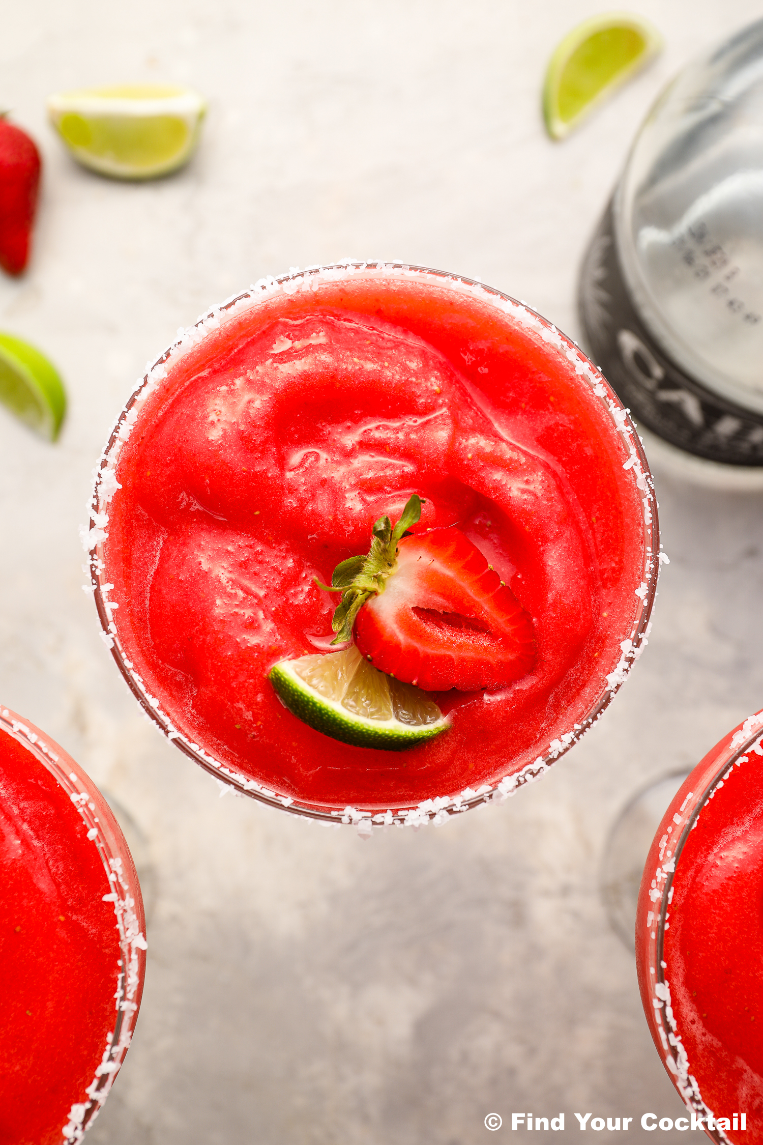 A close-up of a frozen strawberry margarita garnished with a strawberry slice and a lime wedge, with a salted rim and lime wedges in the background.