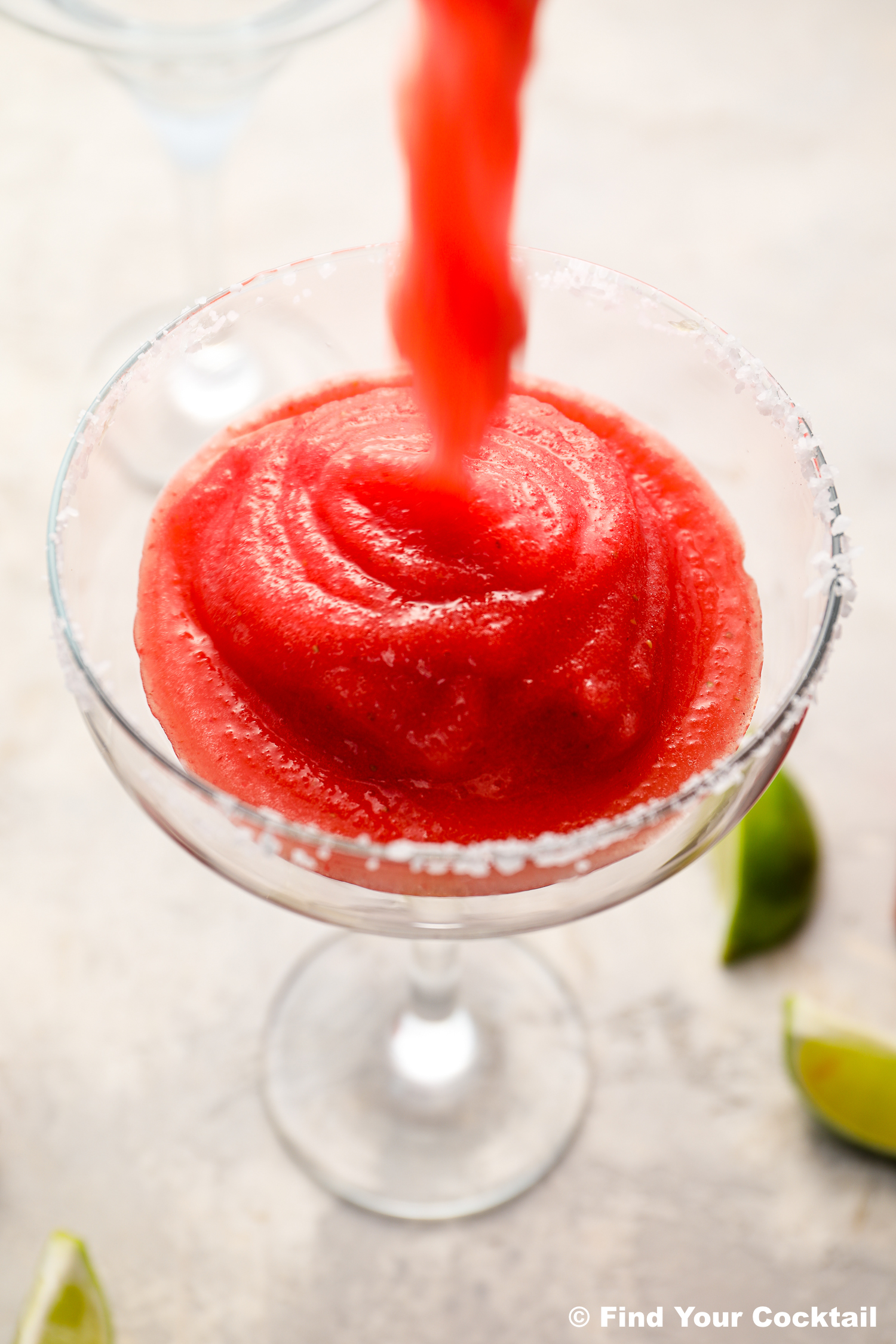 A frozen red cocktail is being poured into a salt-rimmed glass, with lime wedges in the background.
