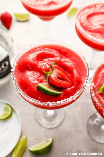 A frozen strawberry margarita in a salt-rimmed glass, garnished with a fresh strawberry slice and lime wedge. Other glasses and lime pieces are visible in the background.