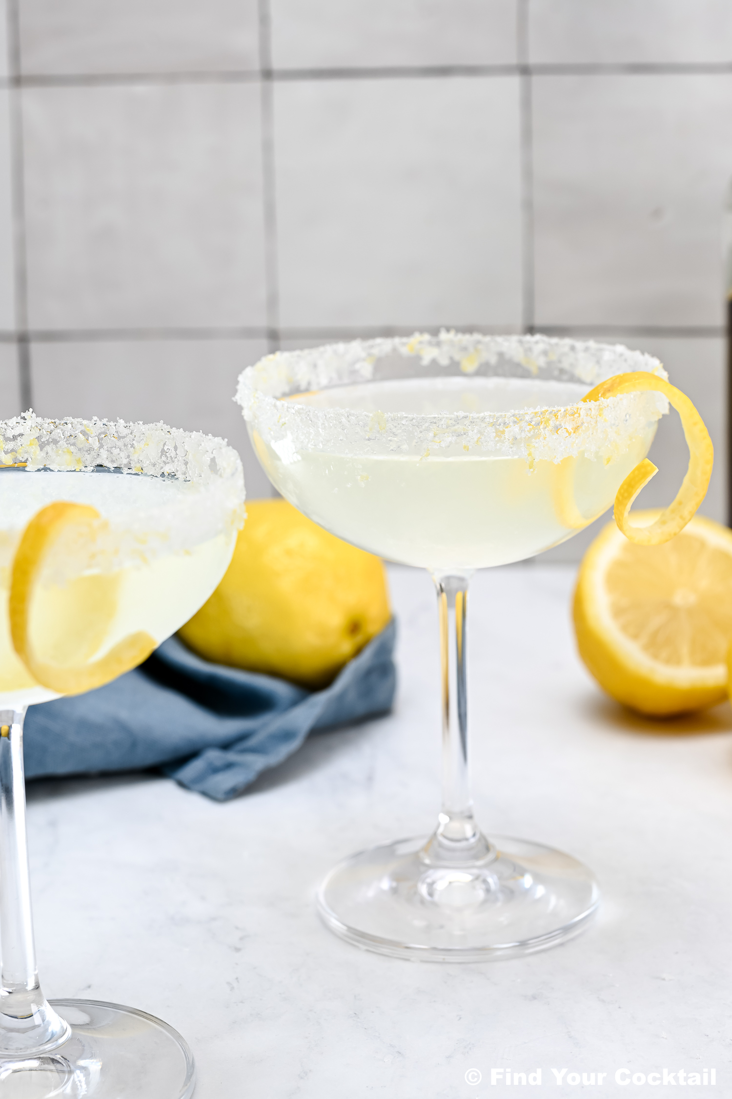 Two coupe glasses with a sugared rim, filled with a pale yellow cocktail, garnished with a lemon twist; lemons and a blue cloth sit in the background.