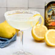 A sugared-rim cocktail glass with a clear drink and lemon twist garnish sits on a white surface with lemons and a bottle in the background.