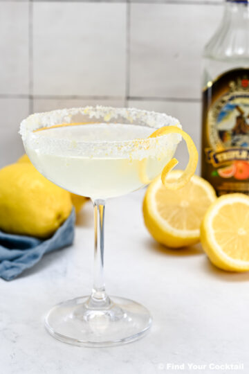 A sugared-rim cocktail glass with a clear drink and lemon twist garnish sits on a white surface with lemons and a bottle in the background.