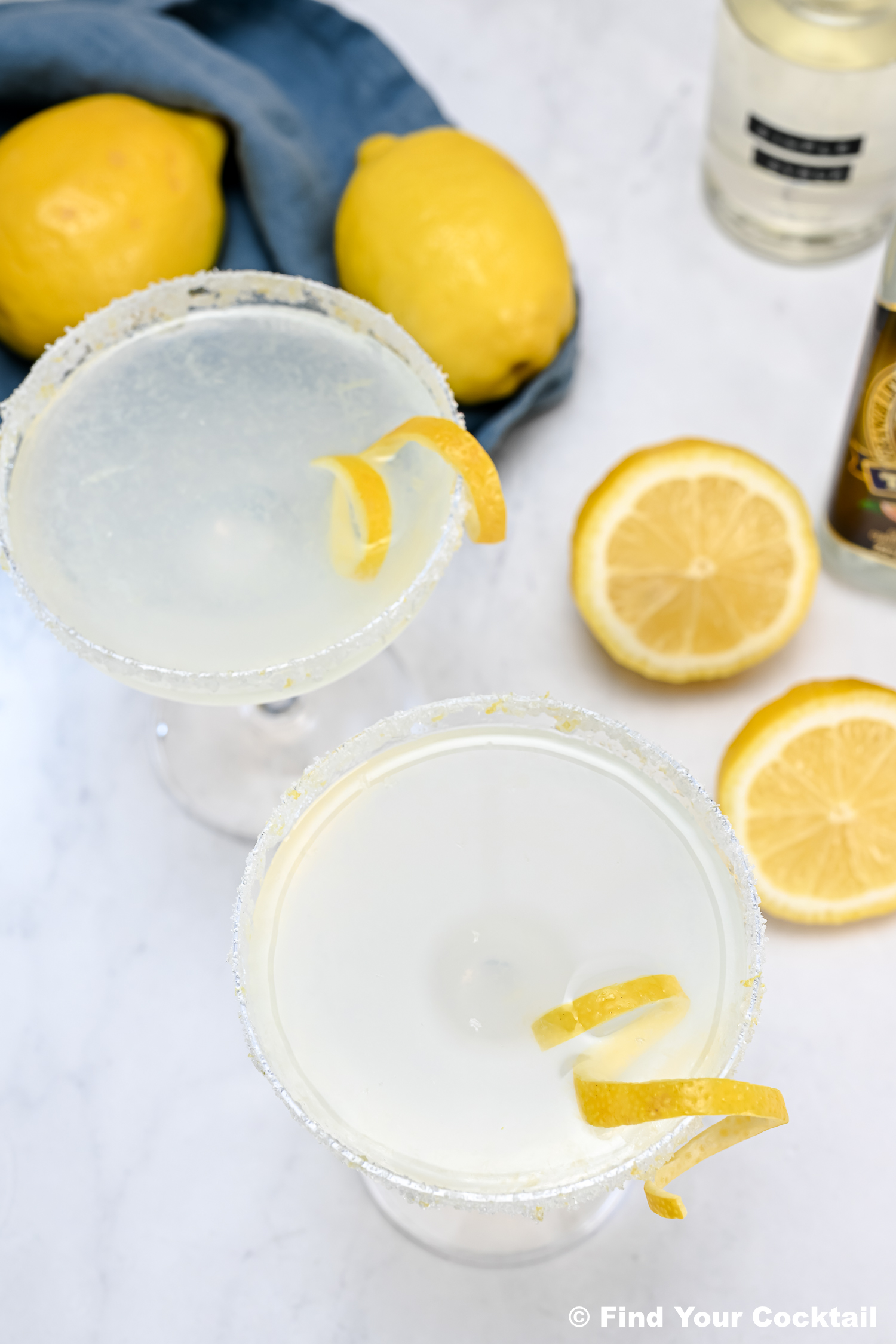 Two cocktails with sugared rims and lemon twists are on a marble surface, surrounded by whole and sliced lemons, a blue cloth, and a bottle.
