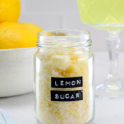 A glass jar labeled Lemon Sugar sits on a table, with a bowl of lemons and a cocktail glass in the background.