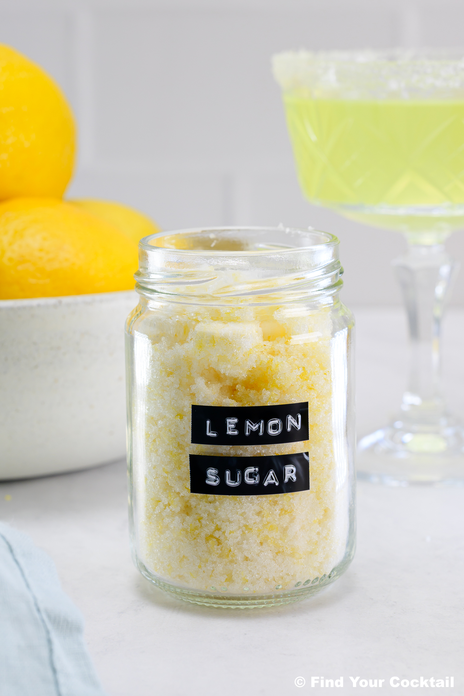 A glass jar labeled Lemon Sugar sits on a table, with a bowl of lemons and a cocktail glass in the background.