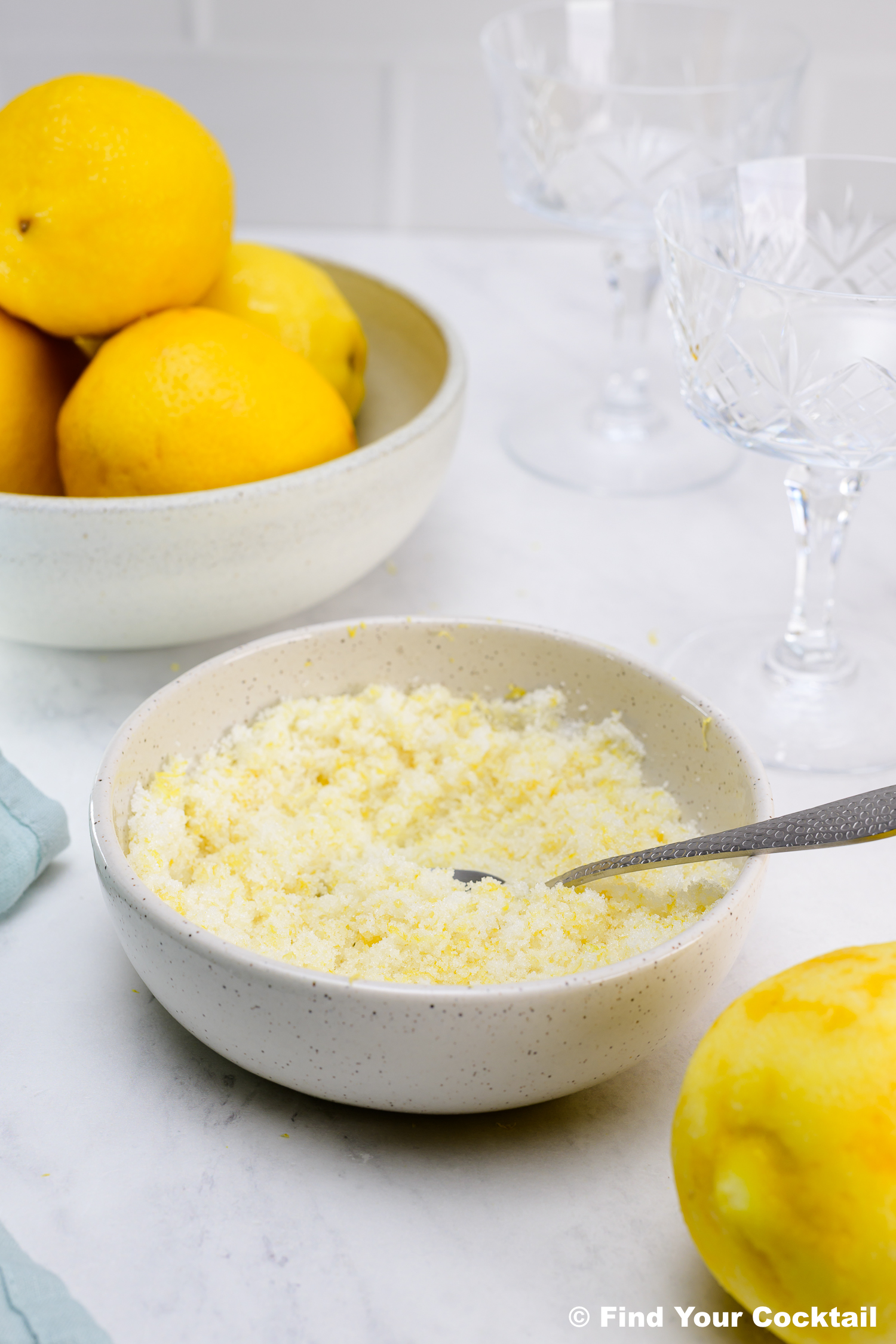 A bowl of grated lemon zest and sugar with a spoon, next to whole lemons in a bowl and empty crystal glasses on a white countertop.