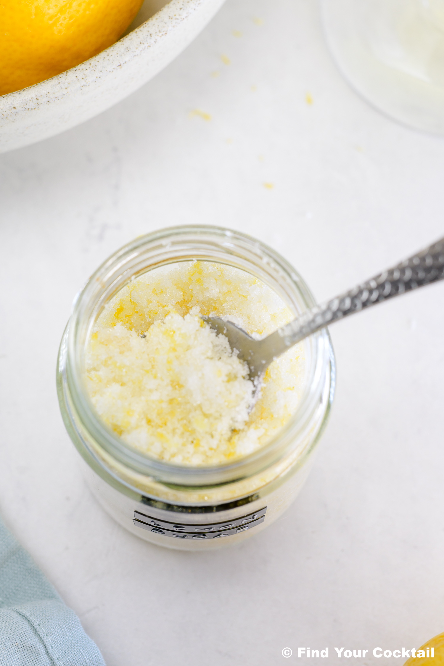 A spoon in a glass jar filled with a granular lemon sugar mixture, with a lemon partially visible in a bowl nearby.