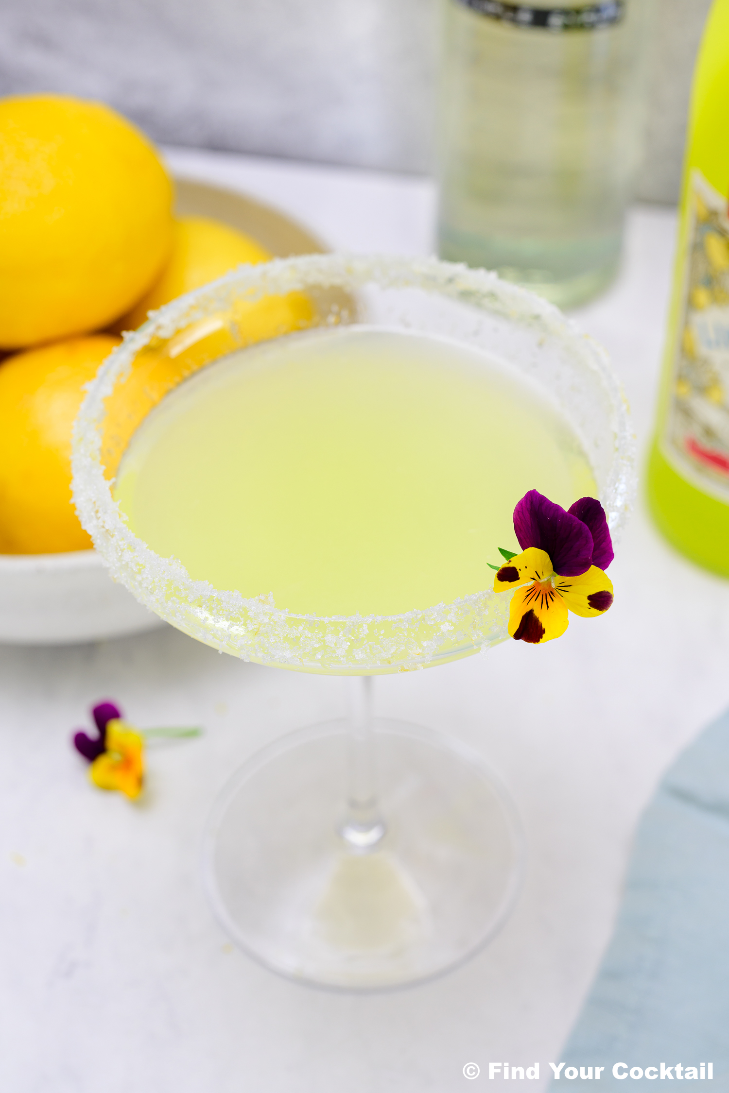 A pale yellow cocktail in a sugared-rim glass, garnished with a small edible flower. Lemons and bottles are visible in the background.