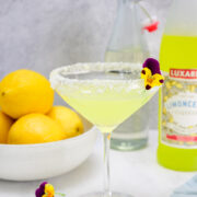 A yellow cocktail in a sugar-rimmed glass garnished with a pansy flower, next to a bowl of lemons, a bottle of limoncello, and a clear bottle of water.