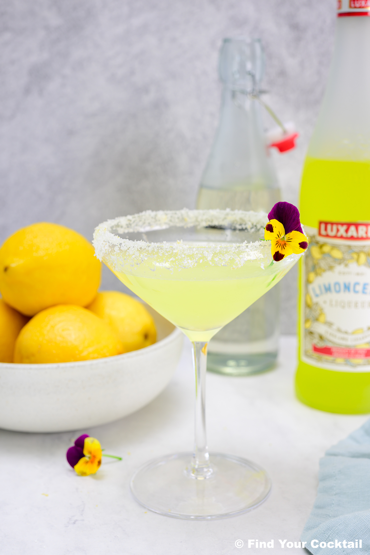 A yellow cocktail in a sugar-rimmed glass garnished with a pansy flower, next to a bowl of lemons, a bottle of limoncello, and a clear bottle of water.