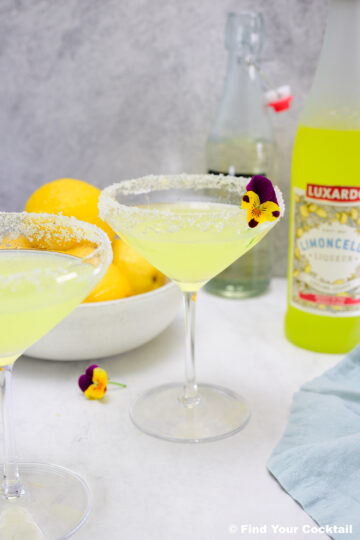 Two cocktails in rimmed glasses garnished with edible flowers, with a bowl of lemons, a bottle of limoncello, and a glass bottle in the background.