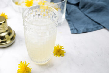 Clear woven glass filled with a pale yellow beverage, surrounded by dandelion flowers on a white surface, with a blue cloth and shaker in the background.