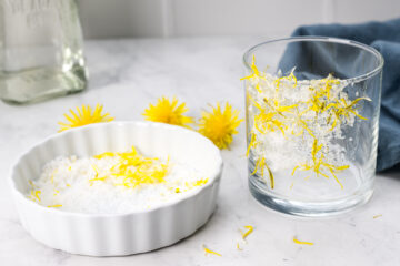 A glass rimmed with a mixture of salt and yellow zest sits beside a white dish containing the same mixture, with dandelion flowers in the background on a marble surface.